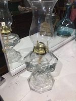 Full view of the vintage glass oil lamp showing the clear glass chimney, brass burner, and hexagonal base on a reflective surface.