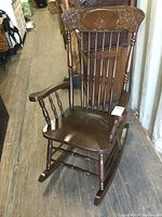 Front angled view of vintage pressed back rocking chair showing carved floral design, spindled backrest and arms, and contoured seat.