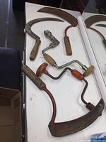 Overview of three sickles with wooden handles and two hand braces on a white countertop showing aged metal blades and wooden handles.