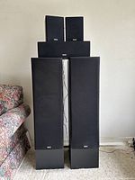 Five Yamaha speakers arranged with two tall floor-standing NS-AP100 speakers at front, a center channel speaker in middle, and two bookshelf speakers on top of center speaker, all with black fabric grilles and Yamaha branding.