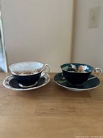 Two vintage bone china cups and saucers on a wooden surface in front of a white wall, showing dark blue and dark teal color sets with floral patterns and gold trimming.