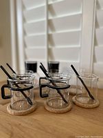 Six vintage Bodum glass coffee cups with black handles and cork bottoms, each paired with a black spoon, displayed on a table with white shutters in background.