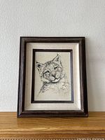 Framed pencil drawing of a bobcat on canvas against a wall, showing the full piece.