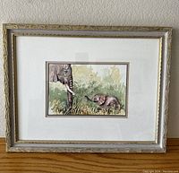 Framed watercolor painting showing two elephants, an adult with tusks and a baby, in nature with grass and foliage. The frame is off-white with gold accents, the painting is matted and behind glass.