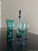 Photo of turquoise blue glass decanter with matching blue stopper and four clear glasses with blue stems and bases arranged on a glass surface under natural light in front of a neutral wall.