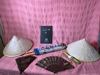 Photo showing all items in the lot including two vintage Asian conical coolie hats, two decorative folding fans, and two umbrellas with Asian motifs laid out on a pink background.