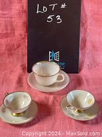 Three coffee cups with saucers: two delicate Bavaria porcelain cups with floral patterns and gold trim, one white Fire-King glass cup with gold trim.