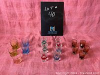 Full front view of all 16 colored liqueur glasses arranged in groups by style against pink fabric background.