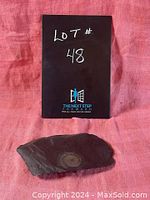 Photo showing the black slate piece with visible ammonite fossil, placed on a pink cloth background along with a black card labeled 'LOT # 48'.