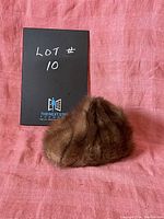 Photo showing the brown fur hat against a pink backdrop with a black card indicating lot number 10, displaying the hat's rounded top and fur texture