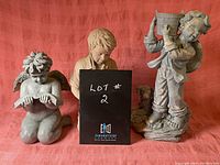 Three lawn ornaments against red backdrop including an angel reading a book, boy kneeling with dog, standing boy with basket.