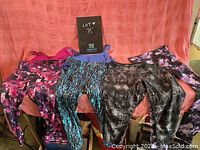 Photo of four pairs of colorful women's workout leggings spread out on a couch, showing various patterns including pink and black, blue and black, black with white streaks, and purple floral designs with two tank tops stacked on top.