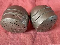 Two vintage modified Volvo metal automotive parts placed side by side on a reddish fabric background, showing perforated ends and textured surface.