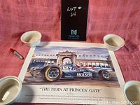 Photo of the unframed poster laid flat between three white ceramic bowls and a red marker, showing the entire image including handwritten lot number on a black board in the background.