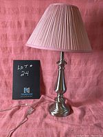 Silver colored table lamp with pleated pink lampshade on a pink cloth background. The lamp is standing upright with its cord visible.