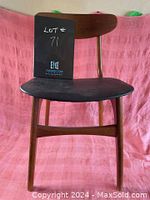 Front view of the teak dining chair showing the wooden backrest and black faux leather seat against a pink fabric background.