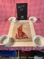 Photo of the unframed poster laid flat with four white ceramic bowls holding edges down, showing detailed view of the colorful Thames Sailing Barge "May" with red sails on water, cloudy sky.