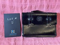 Front view of the black leather briefcase placed on a pink surface, showing the lot number card next to it.