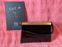 Front view of the glossy black handbag with leather strap and metal clasp closure, placed against a pink backdrop.