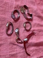 Six vintage watches displayed on pink fabric, showing a variety of metal bands and one with a red leather strap, different watch face shapes and sizes.