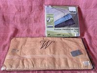 Photo showing a vintage light brown/beige towel with embroidered 'W' laid flat, alongside the packaged Tukan branded towel set.