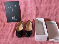 Pair of black women's slip-on shoes size 9.5 with leather detail displayed next to an open white shoebox on a pink cloth background.