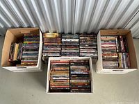 Photo showing three boxes filled with a large number of DVDs stacked inside, placed against a storage unit wall.