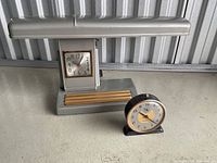 Two retro clocks displayed together on a table with a corrugated metal background. One small round clock in front, one desk lamp clock behind.