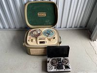 Open Saja reel-to-reel tape recorder showing two reels of tape, control knobs, and speaker grill inside the beige case with green felt lid featuring Saja logo.