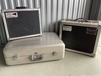 Three equipment cases lined up against a metal wall inside a storage unit: black Audio Hardware Inc. case, beige hard-shell case, and black Tuf Case Plus case with Music Mart label.