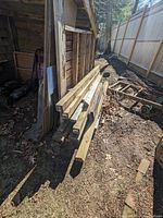 Four 10-foot 4x4 pressure treated lumber pieces stacked on ground leaning against a wooden shed. Visible dirt and wear but intact.
