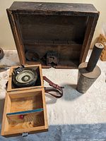 Antique wooden cash drawer with internal metal lock and finger pulls, handheld compass in wooden box, and heavy steel conical tool resting on table.