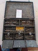 Open metal case showing multiple compartments filled with various drill bits organized by size and type.