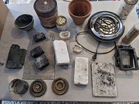 Top-down view of various casting materials including charcoal blocks, casting sand canister, crucibles, rubber bowl, fire bricks, metal hotplate, green wax mold, and terracotta pot.