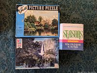 Three vintage cardboard jigsaw puzzles displayed, two sealed including sealed Milton Bradley Yosemite Falls (500 pieces) and 1974 Seasons Calendar, and one used Guild Millpond puzzle.