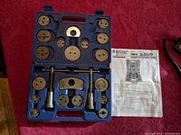 Complete blue carrying case with 18 metal pieces fitting in molded slots and instruction sheet beside it on red cloth surface.