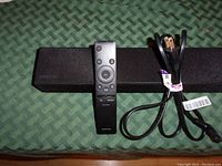 Front view showing black Samsung soundbar with fabric grille, Samsung branding visible, black remote control placed on top, and coiled power cord beside it.