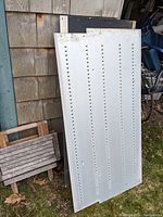 Aluminum sheets leaning against a wooden exterior wall, one sheet with multiple perforated rows visible.