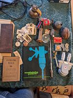 Overview of the lot showing books, tubes, brass items, a candle holder, photo cards, and other metal parts on a patterned tablecloth.