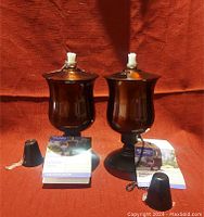 Two dark metallic outdoor tabletop torches with white fiberglass wicks, black snuffers, tags attached, placed against a reddish fabric background.