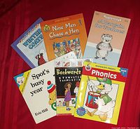 Six children’s early reader books arranged on a red surface, front covers visible showing titles and illustrations including cartoon animals and colorful designs.