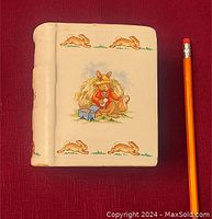 Front view of the Bunnykins ceramic piggy bank shaped like a book with a bunny illustration playing a musical instrument and smaller bunny images surrounding it, set next to a pencil for scale.