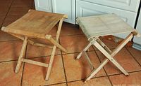 Two 1980s wooden fold-up camping stools with fabric seats on tiled floor, showing natural wood finish and beige fabric.