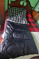 Sleeping bag laid open on a bed showing black exterior and plaid flannel interior, illustrating size and padding.