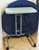Hospital bed table folded with blue chair behind. Shows frame and caster wheels.