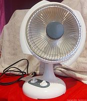 Front view of small white electric radiant heater on pedestal base with protective metal grille covering heating element and power cord attached.