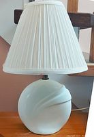 Photo of white porcelain table lamp with pleated shade on wooden surface against a wall.