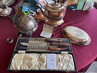 Photo of all items together on table: silver hinged kettle with stand, covered serving dish, George Butler & Co cutlery set in open case, napkin rings.