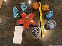 Photo showing the full lot of nine assorted art glass pieces including a large orange star-shaped paperweight, two blue-tinted owls, black heart-shaped piece, and several spherical paperweights.