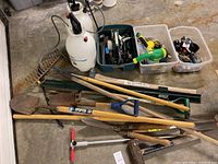 Photo showing large assortment of gardening tools laid on concrete floor including shovels, rakes, sprayers, garden stakes, and hand tools in tubs